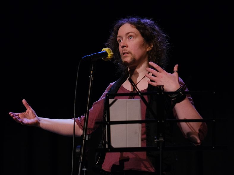 A slender, white, goatee bearded person with curly, shoulder-length heair of an indeterminate colour is standing, spotlit, gesturing broadly on a darkened stage behind a microphone and a basic music stand with an ereader on it. They give the impression that they're expanding on a difficult point.