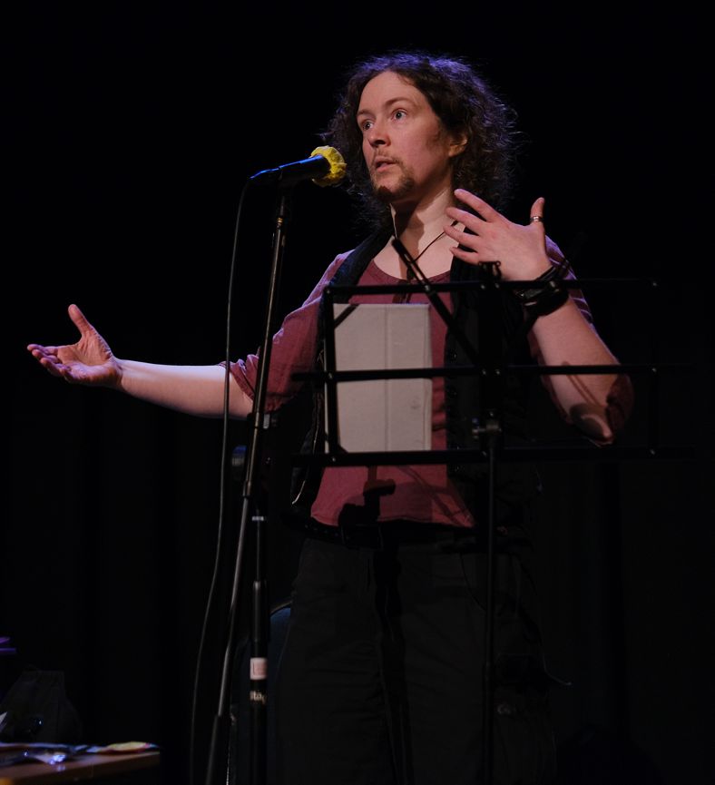 A slender, white, goatee bearded person with curly, shoulder-length heair of an indeterminate colour is standing, spotlit, gesturing broadly on a darkened stage behind a microphone and a basic music stand with an ereader on it. They give the impression that they're expanding on a difficult point.