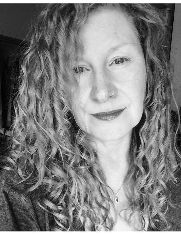 A close-up, black-and-white shot of a pale, white woman in early middle-age with long, blonde, very curly hair and freckles across her nose and cheekbones. She is wearing a small jewel on a delicate chain around her neck, a small, light metal nosering, and subtle makeup. Her smile is faint and her eyes kind.