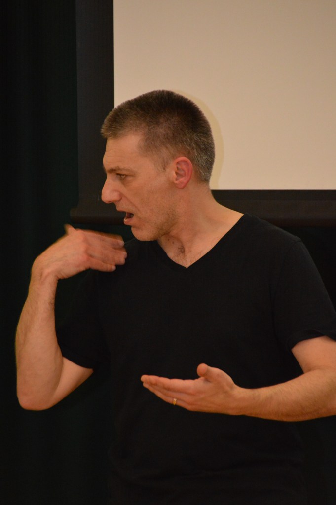 a slender, middle-aged white man with a slightly ruddy complexion and light stubble has his head turned to one side, presenting a strong profile in front of a large projection screen, mouth open as he gestures emphatically, one hand, with a wedding band, on one finger, cupped in front of him, the other arm crooked high as he beckons towards his shoulder. He has very short-cropped, salt-and-pepper hair, and is wearing a black teeshirt. End image description.