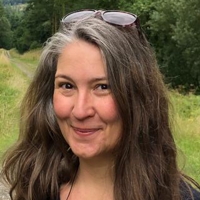 Headshot of a white, tanned woman in early middle age smirks mischievously at the camera against a backdrop of woodland and meadow on a slightly overcast day. She has long, mid-brown hair with a hint of auburn, growing in silver at the roots and temples and happy, dark brown eyes. Her sunglasses are perched on top of her head.