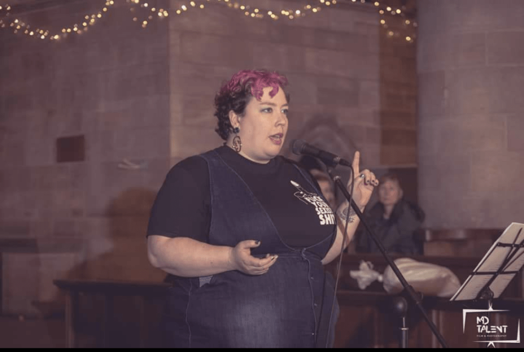 A performance shot of a fat, white person with short, curly, mid-brown hair dyed bright magenta on top is standing behind a microphone and music stand in what appears to be a large, old church. They are immaculately made-up with large hoop earrings and metallic nail polish, gesturing authortitatively, one finger raised, the other hand cupped beside them. They are wearing a dark, navy blue pinafore dress over a black teeshirt which bears the caption "Fuck Your Sexist Shit", and have a tattoo on the wrist of the hand pointing upwards.