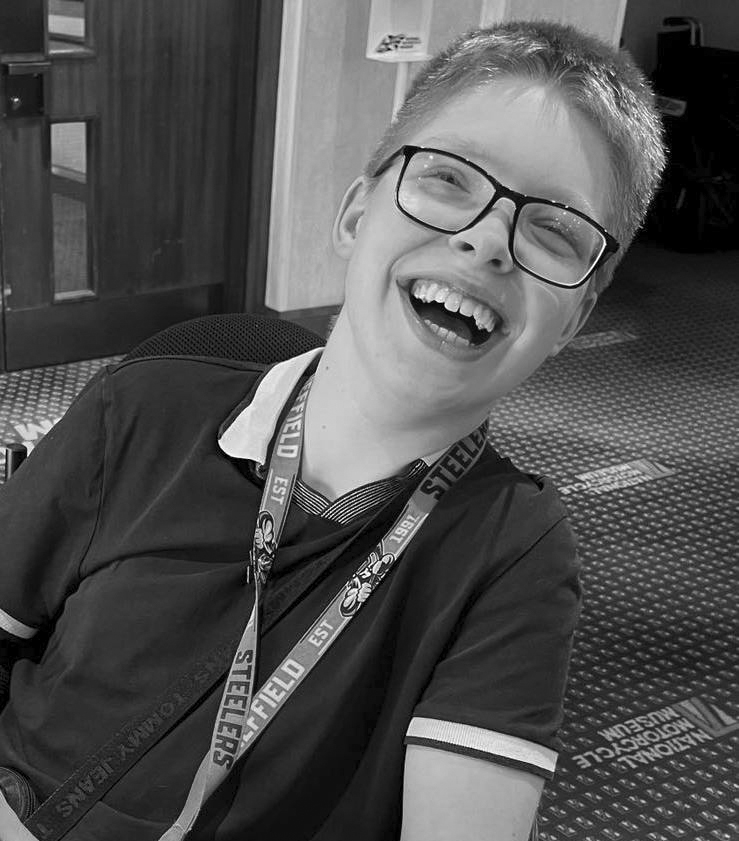 A close-up, black-and-white shot of a pale, white young man with very short, blonde hair and heavy-rimmed, square glasses. He is leaning back at a slant in his wheelchair and laughing heartily, blue eyes very nearly shut in mirth, grin huge and open. He is indoors at the National Motorcycle Museum, according to the logo on the carpet behind him, and is wearing a dark polo shirt, short-sleeved, with white collar and cuffs, and a Sheffield Steelers lanyard around his neck.