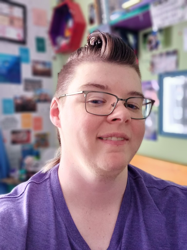 A young, white person with a round face, slightly ruddy complexion, and dark-rimmed, square glasses tilts their head back, smiling wryly at the viewer. They have light stubble on their chin and throat, and pinkish hair swept up in a slicked-back quiff with a side-shave. They are wearing a loose, purple, v-necked top. The indoor walls behind them are crowded with images, framed and unframed, all out of focus.