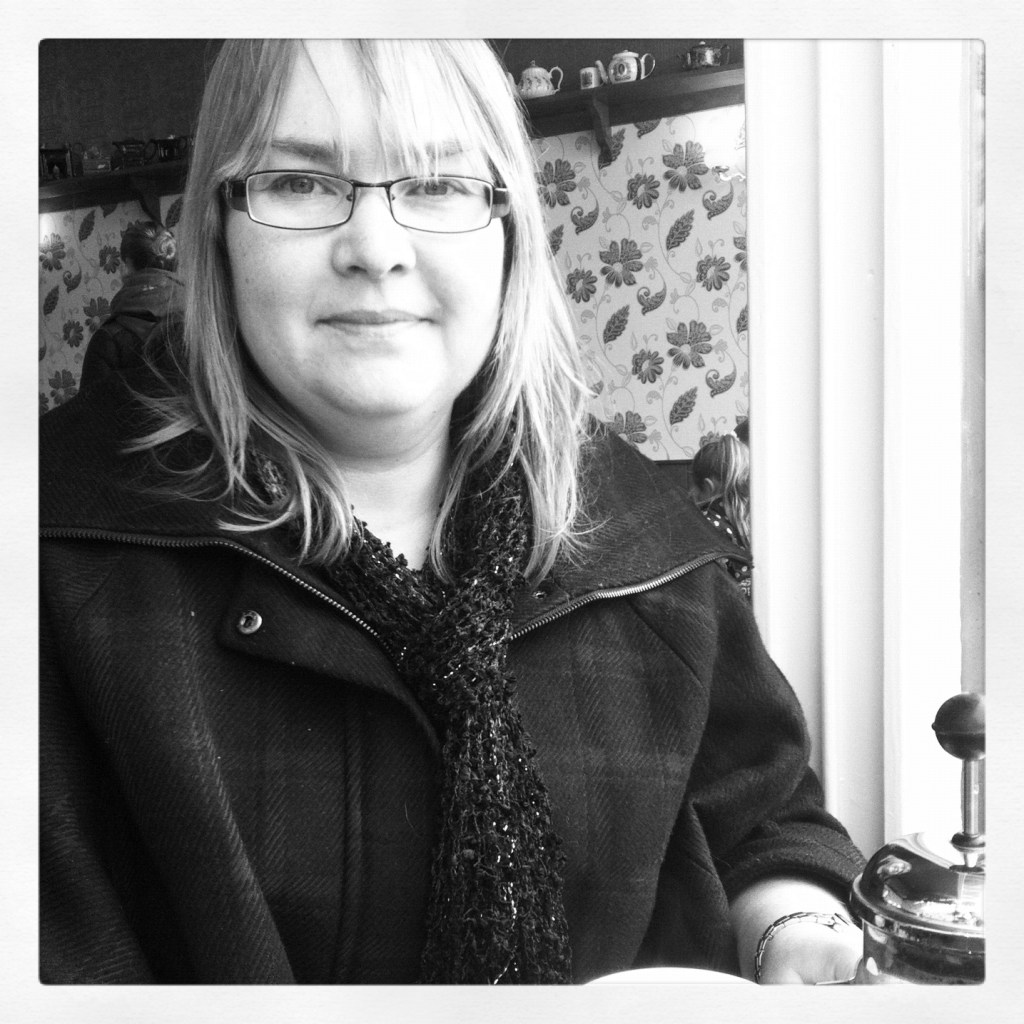 a black-and-white shot of the upper body of a pale, round-faced, white woman with shoulder-length, straight, very pale hair, sitting at a table in a coffee shop, next to a window. She is still wearing a dark, subtly plaid coat, zipped up, with a broad collar splayed to show a slightly sparkly, dark, loose-knit scarf. Her smile is small and complex and she gazes directly at the camera with calm, light eyes through rectagular glasses with light, metallic frames, a coffee pot at one hand.