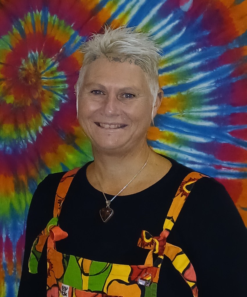 a white woman with a round face and short, spiky, silver-white hair is grinning at the camera. She has multiple face and ear piercings, and tattooes of moon symbols along her hairline at the top of her forehead. She is wearing bright, multi-coloured dungarees over a black, long-sleeved top. Behind her is a rainbow-coloured, tie-dye sheet.