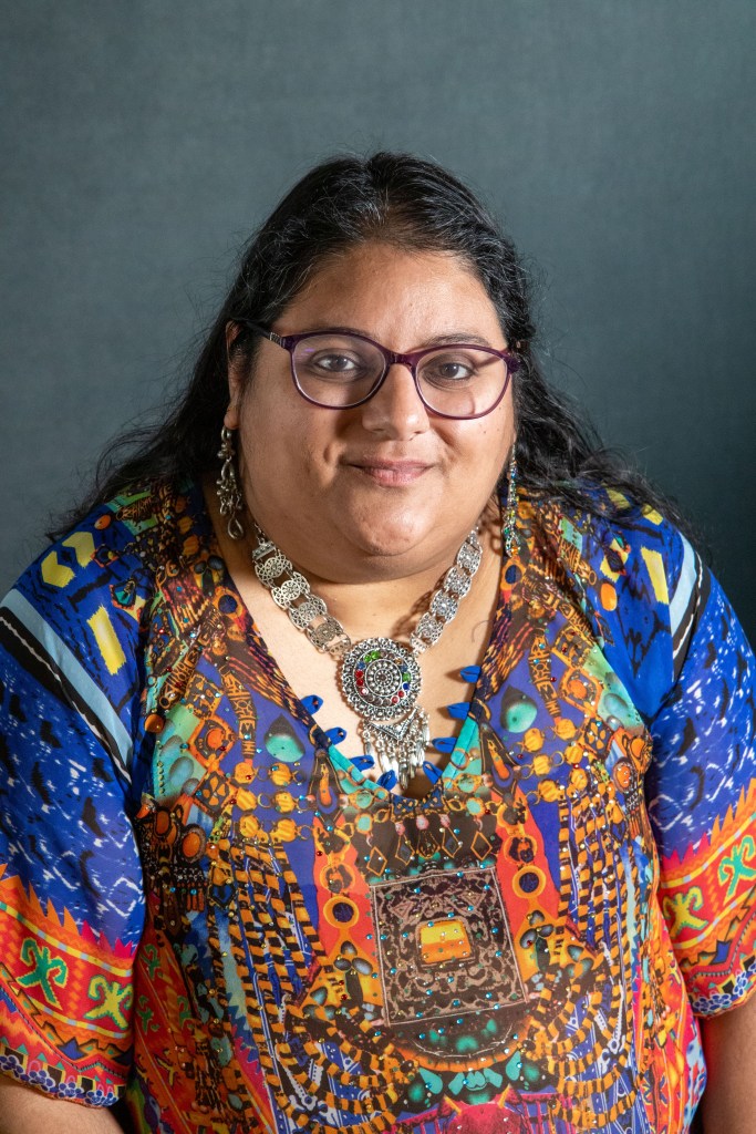 A brown skinned woman with glasses wearing a colorful top with a warm smile on her face