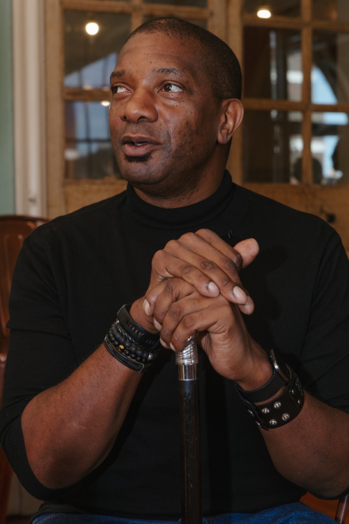 Rick, a black, clean-shaven man with very short-cropped, black hair, is seated dressed in a black polo neck blue jeans and balck leather bracelets and cuffs. His sleeves are rolled up to the elbow. He is looking off camera to his left listening to someone out of frame. His hands are resting on the top of his cane with right hand over left.