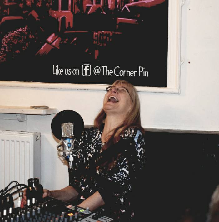 Rose, a white middle-aged female, is sitting behind a table with a large radio mic on it and has thrown her head back in laughter. Her ever-present flask of hot tea is at her side.
