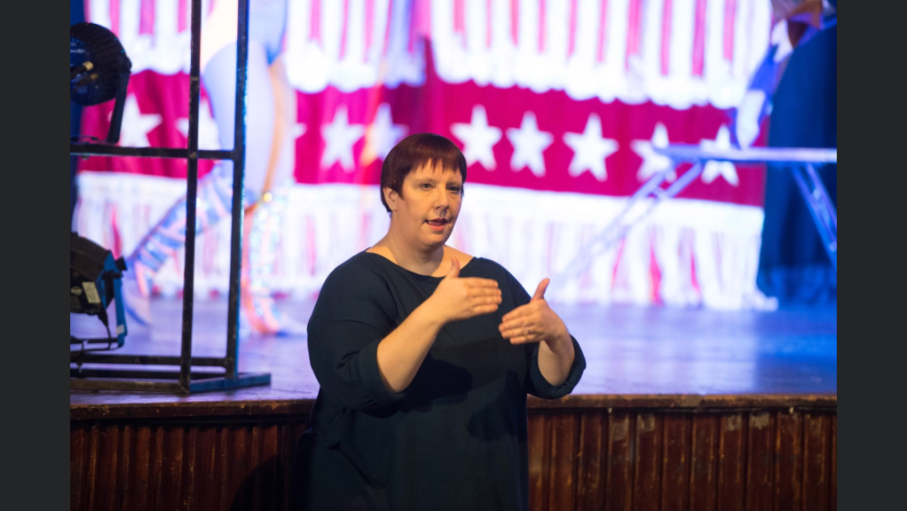 a white, round-faced woman with straight, very short, glossy, reddish-brown hair is gesturing with a very focused expression on her face, mouth slightly open, wearing a plain, loose, dark blue top with three-quarter sleeves. Behind her is dark, wooden stage with the legs of a very glamorous performer in a leotard and sparkly boots against a red-and-white, star-spangled banner, clearly about to do something very acrobatic.