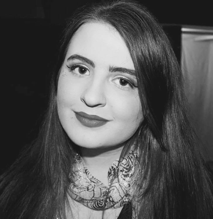 This is a black and white photograph of Sylvia. She is a caucasion female, with dark eyes and hair, wearing dark lipstick, and classic makeup consisting of winged eyeliner and soft features. Her hair is straight and shiny. She wears a dress scarf around her neck and is looking straight forwards at the camera with a slight smile.